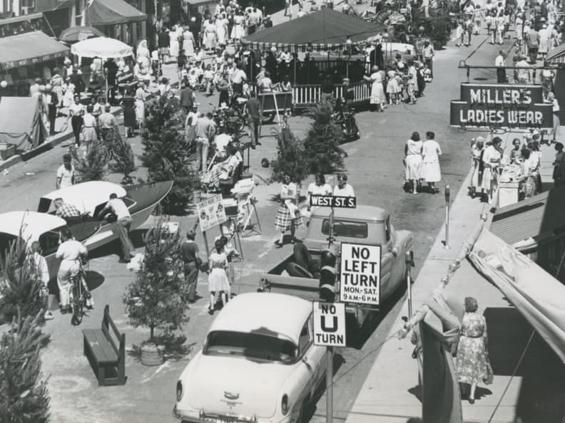 Photo en noir et blanc de la rue Mississaga, à l’intersection de la rue West, montrant une foule de clients et de marchands, plusieurs arbres, et une enseigne où est écrit « Miller’s Ladies Wear ».