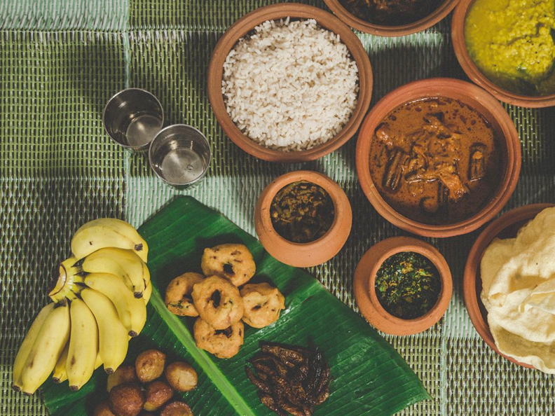 Une table sur laquelle sont étalés divers plats traditionnels, avec du riz, des currys et des bananes sur une natte tissée verte. Les plats sont servis dans des pots en terre cuite.