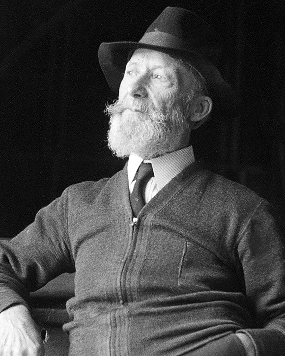 Photographie en noir et blanc d’Ozias Leduc, assis, âgé de 70 à 75 ans. Il porte un chapeau et porte la barbe. Il a la main gauche dans sa poche de pantalon.