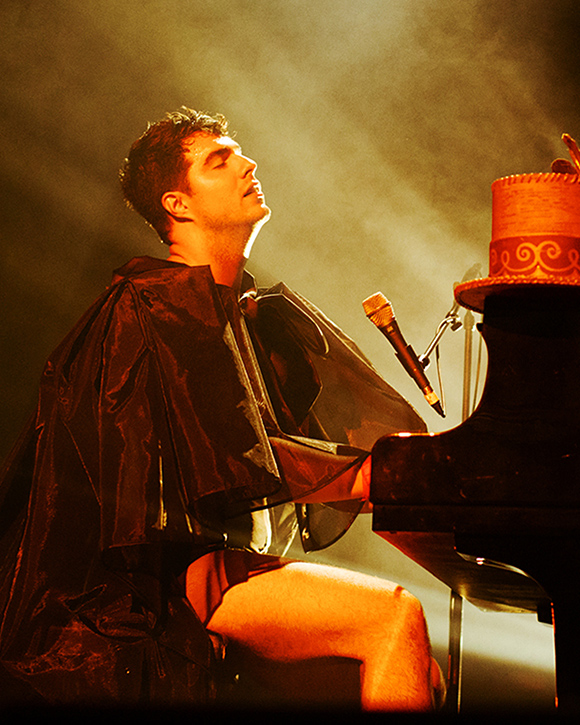 Un homme vêtu d’une cape est assis au piano. Son chapeau haut de forme est posé sur le piano. Ses yeux sont fermés.