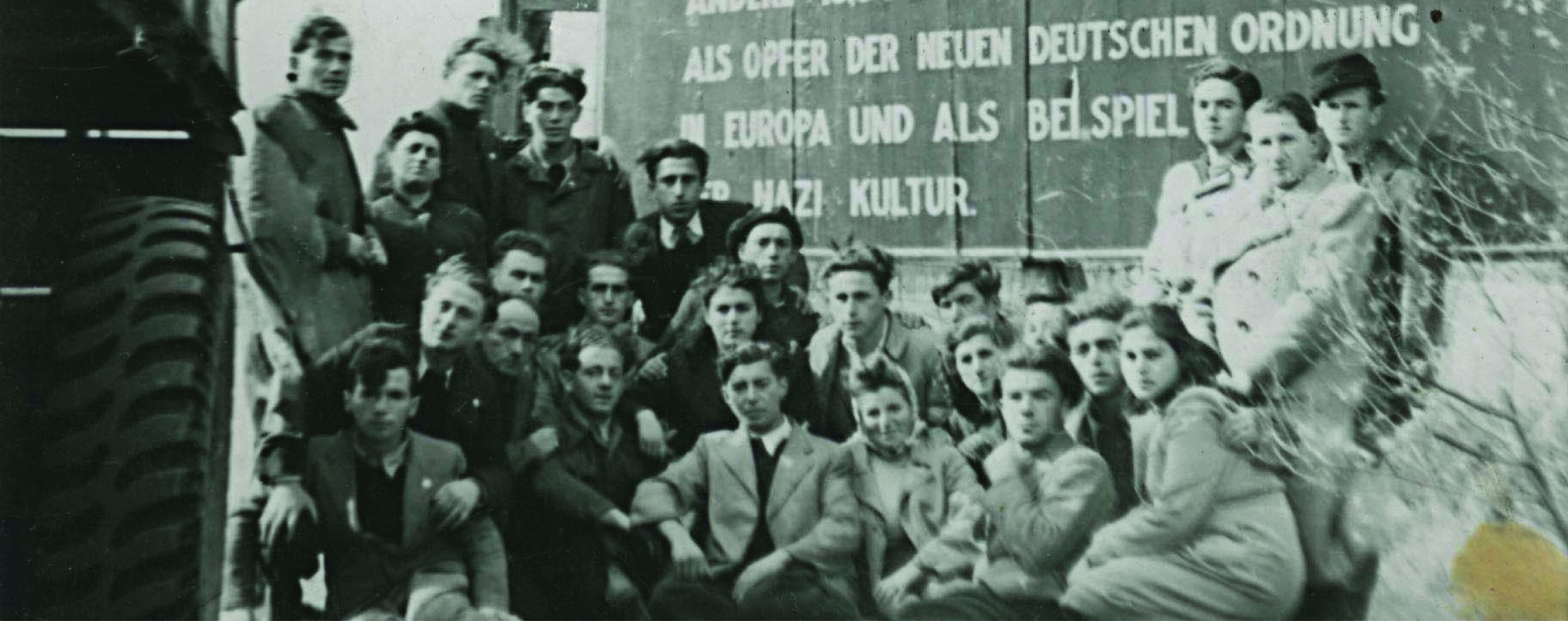 Photo en noir et blanc : Un groupe d'hommes et de femmes posant devant un panneau en allemand marquant l'entrée du camp de personnes déplacées de Bergen-Belsen.