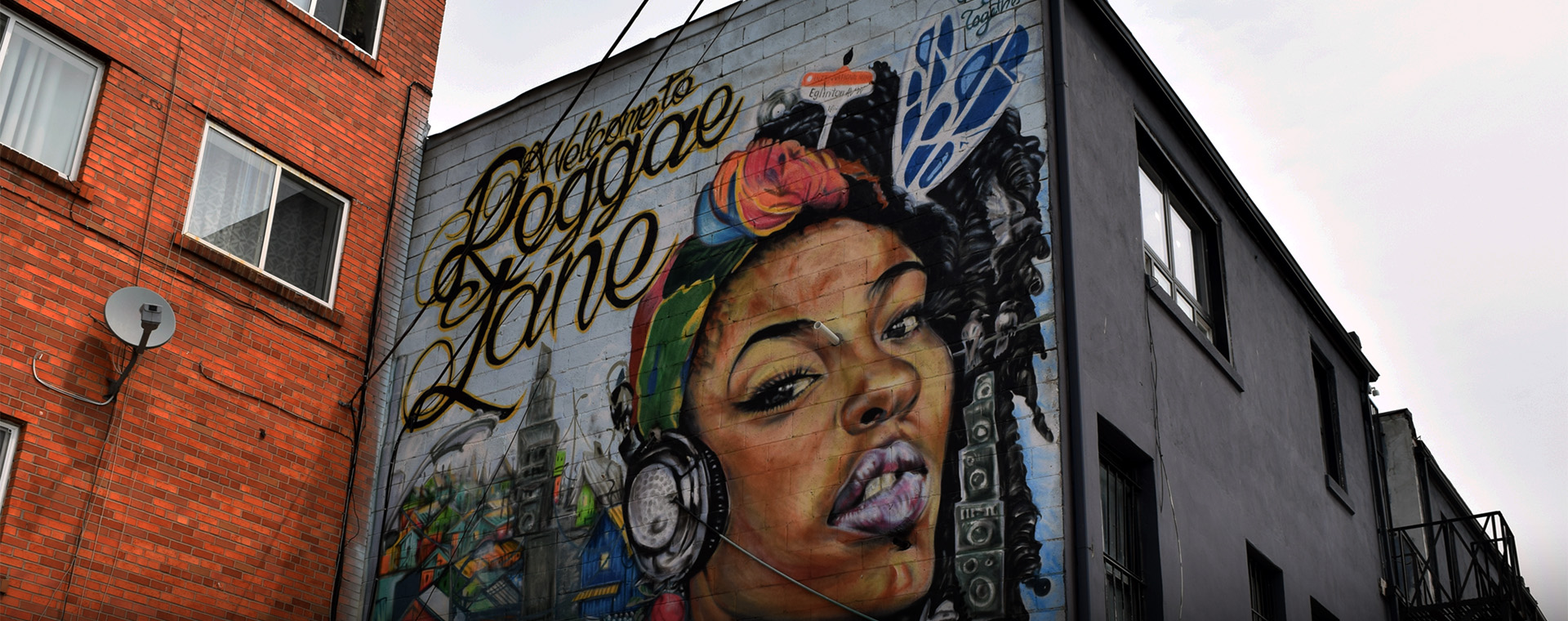 Une peinture murale de Reggae Lane, à Toronto, intitulée « Bienvenue à Reggae Lane ». Le portrait d’une jeune Jamaïcaine portant un casque d’écoute y est dépeint; elle regarde vers le spectateur, et est entourée d’illustrations de drapeaux jamaïcains et de lieux communautaires.