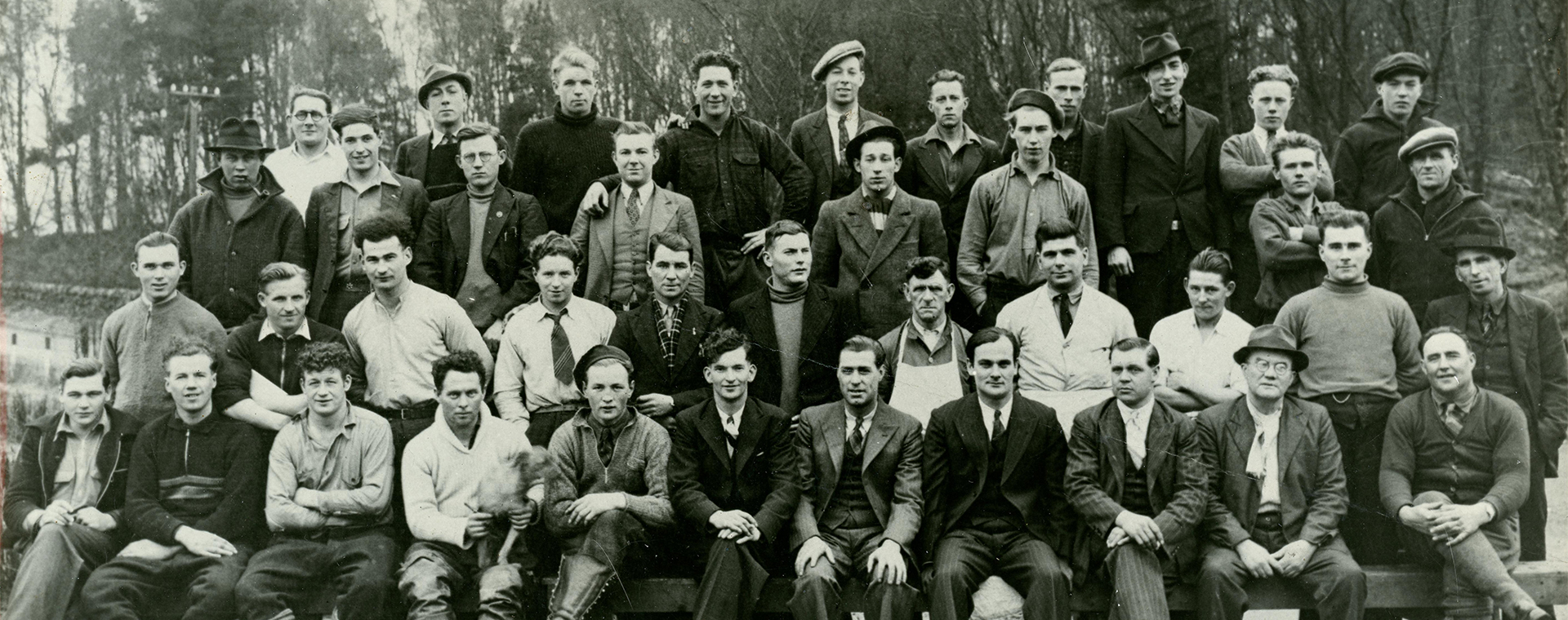 Photographie en noir et blanc montrant 40 hommes, habillés de façon décontractée ou en costume, posant à l’extérieur en 4 rangées; plusieurs sourient. Une rangée d’arbres se dessine au loin derrière eux.