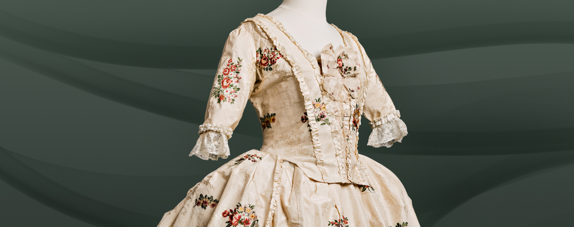 Une rôbe de soirée avec un corsage ajusté au devant et en arrière, démarquée avec une couture, construit du soie, décorée avec des fleurs, en devant d’un fond blanc